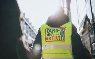 Das Bild zeigt einen Mann von hinten. Er trägt eine neongelbe Streikweste auf der "Tarif gibt's nur aktiv" steht. 