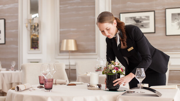 Eine Frau in einem dunkelblauen Kostüm richtet einen Tisch in einem Hotelrestaurant her. Der Tisch ist fein gedeckt mit einer weißen Tischdecke und Blumen.