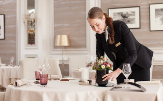 Eine Frau in einem dunkelblauen Kostüm richtet einen Tisch in einem Hotelrestaurant her. Der Tisch ist fein gedeckt mit einer weißen Tischdecke und Blumen. 